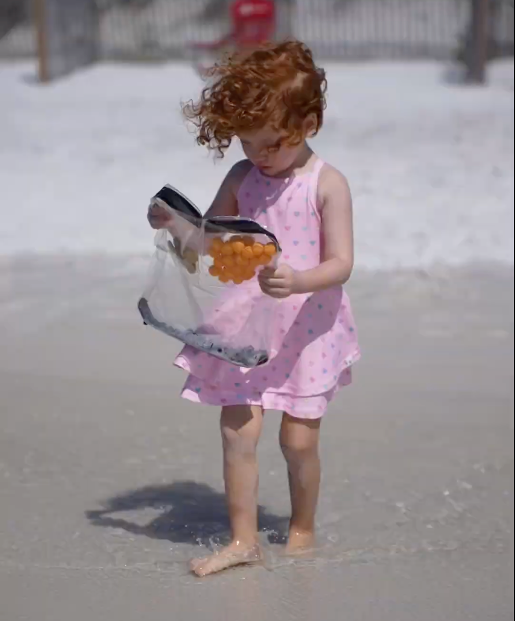 Young girl in pink dress on the beach happily reaching into a clear DivideNPac food-grade organizer bag filled with cheese puffs