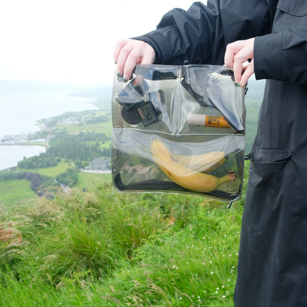 Hands holding a clear DivideNPac food-grade organizer with a banana, on a hillside above a coastal view.