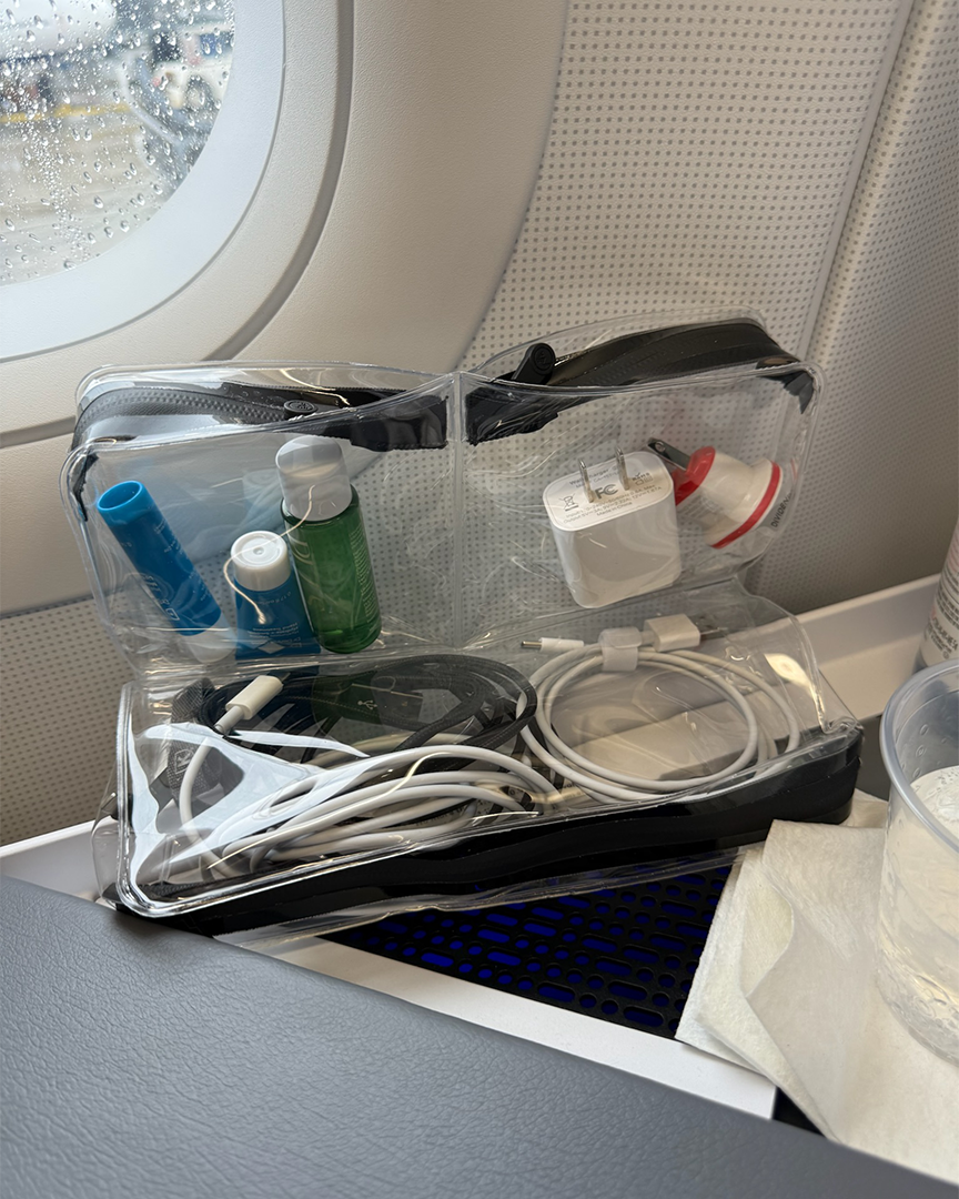 DivideNPac clear organizer on an airplane tray table with chargers, cables, and travel essentials, window seat with rain on the glass.