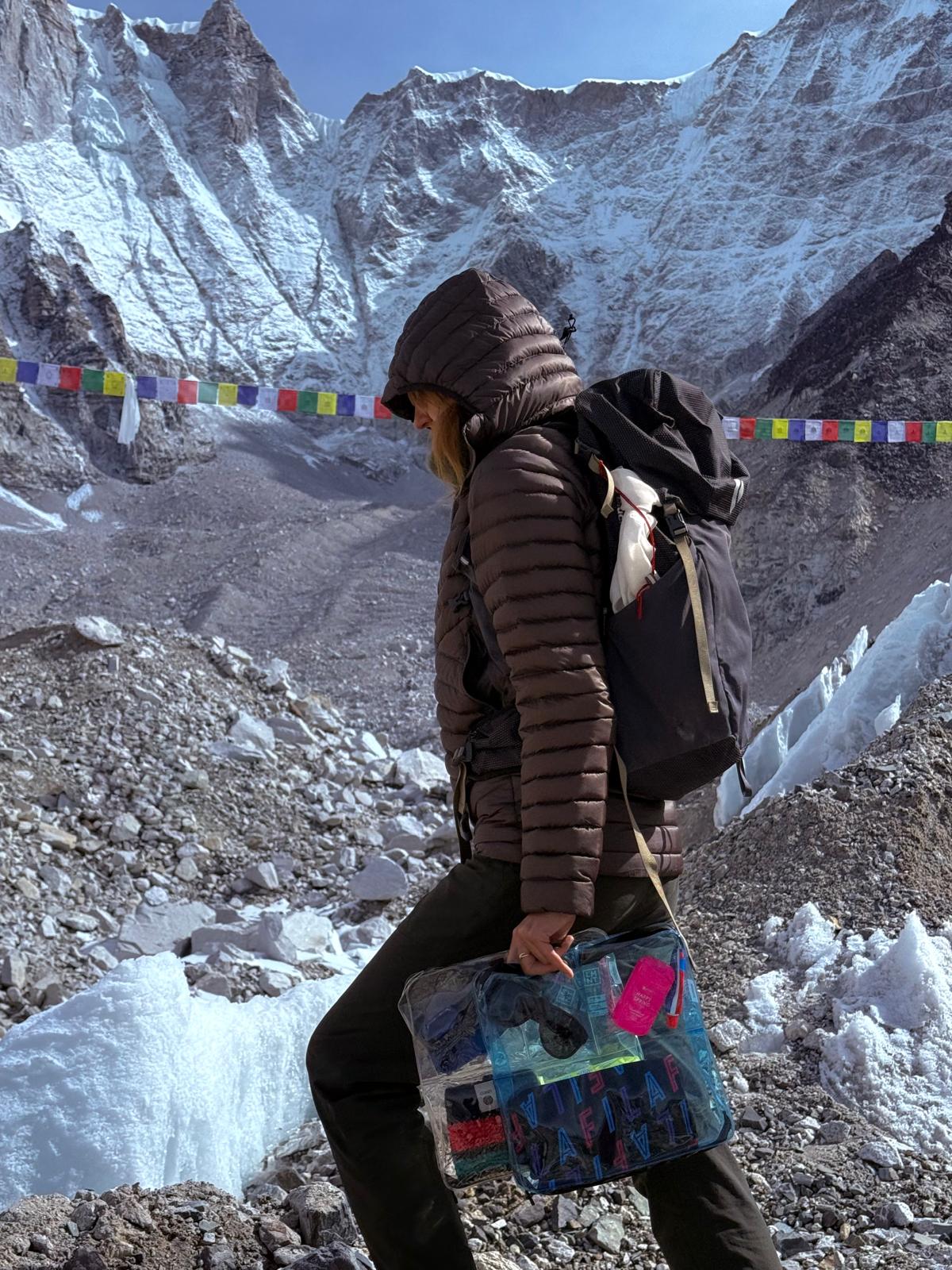 Woman holding a Blue DivideNPac organizer at Everest base camp with Buddhist prayer flags and snowy peaks in the background.