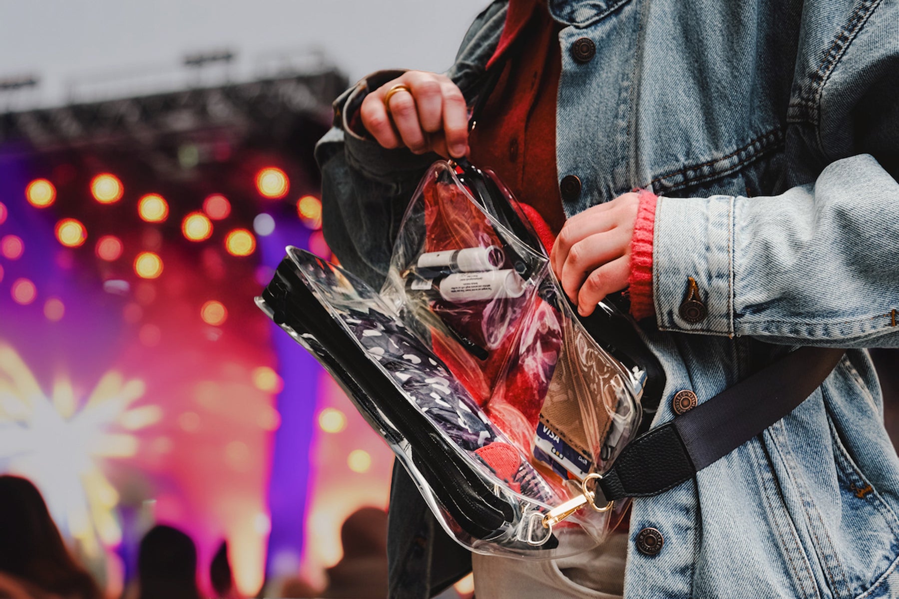 Hand opening a DivideNPac Clear Stadium Bag with three zippered compartments at a concert with stage lights.