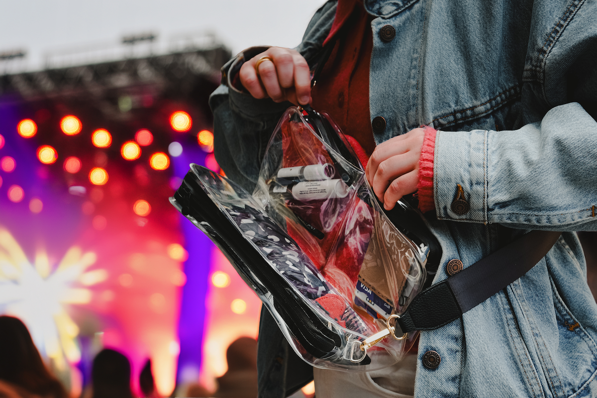 Hand opening a DivideNPac Clear Stadium Bag with three zippered compartments at a concert with stage lights.